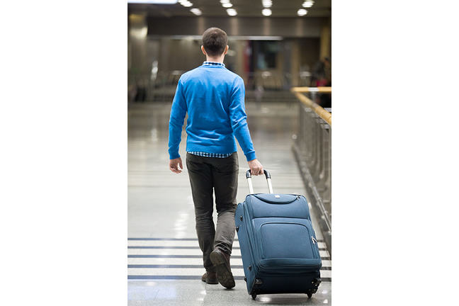 Photo of a man from behind, he’s walking in an airport while pulling his luggage Behind him