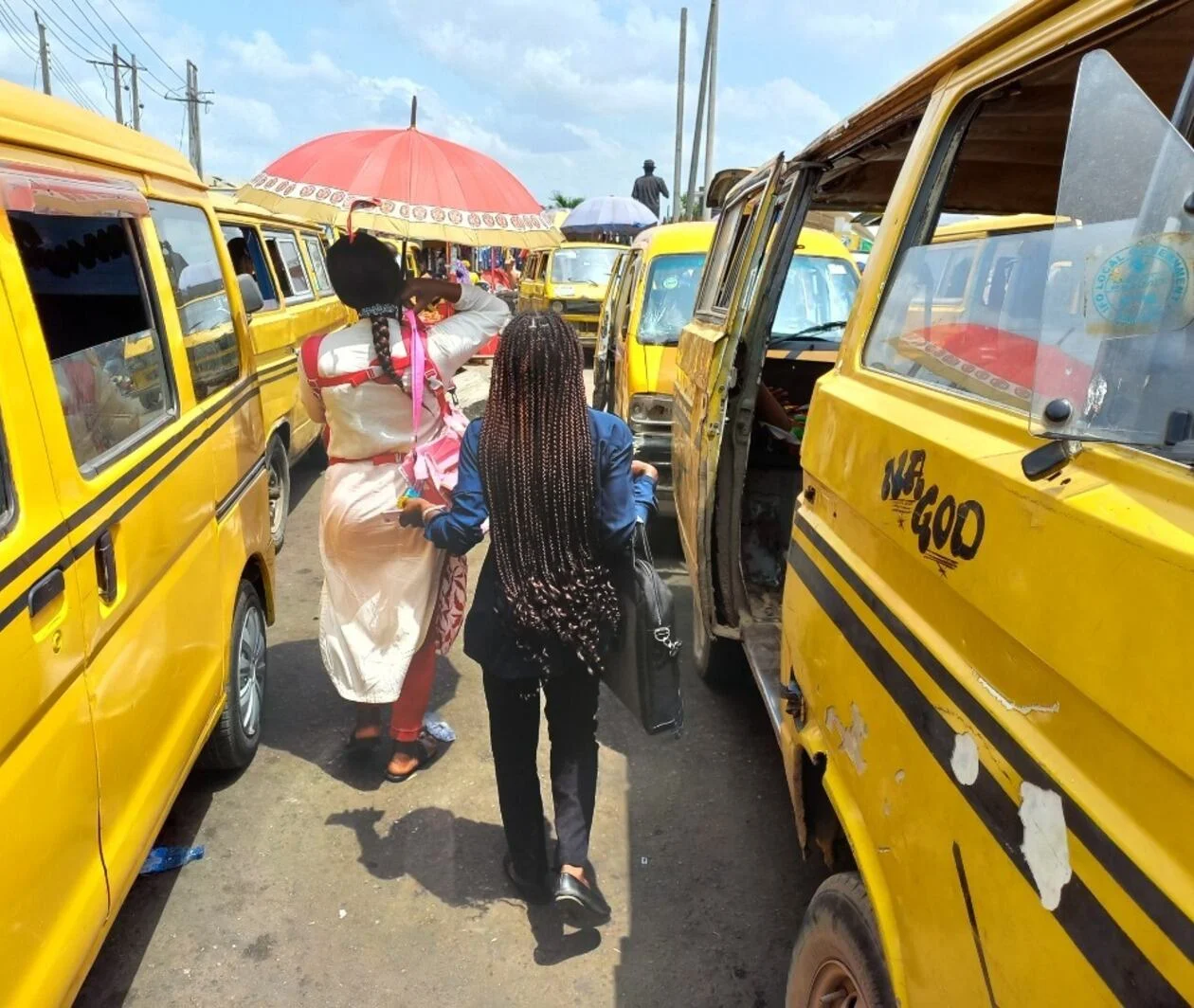 Two women in Lagos, Nigeria, walking between yellow popular (also called informal) minibuses that are parked.