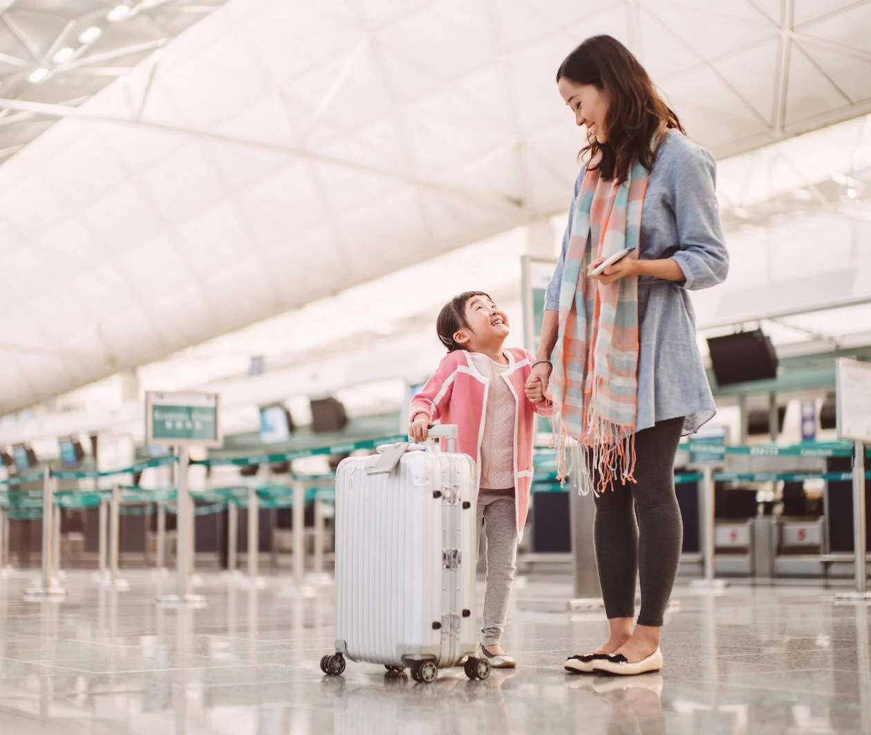 A Chinese mother and her young daughter waiting in an airport, with a silver suitcase ready to begin their travels 