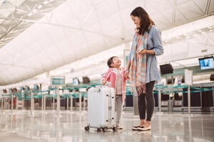 A Chinese mother and her young daughter waiting in an airport, with a silver suitcase ready to begin their travels 