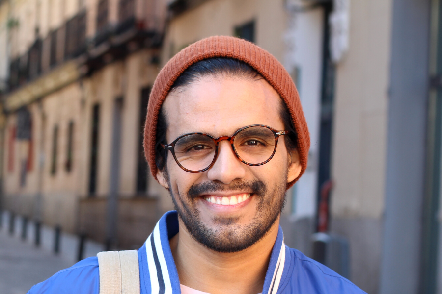 Smiling man wearing glasses, brown beanie, blue jacket and carrying a backpack.