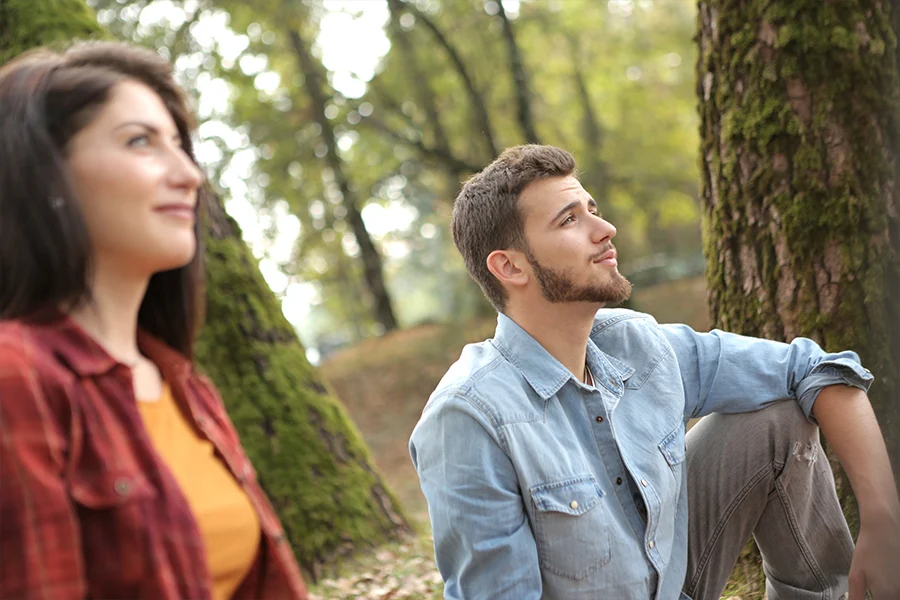 A man and a woman sitting down in the forest looking at something together while smiling.