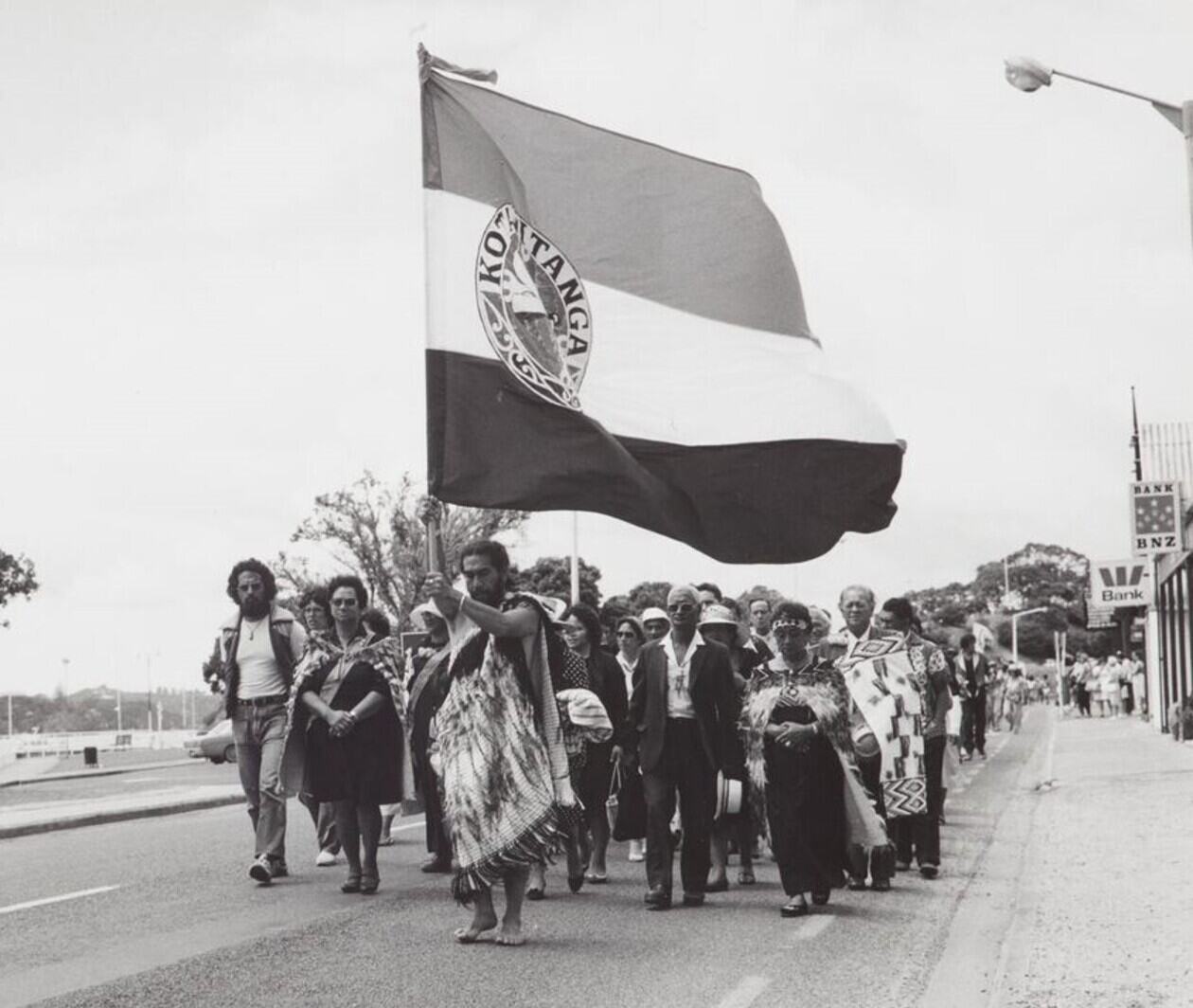 A black and white photograph of Māori men and women. one man and women are in traditional Māori dress, the man is holding a flag. They are in a crowd and are looking down, in prayer.