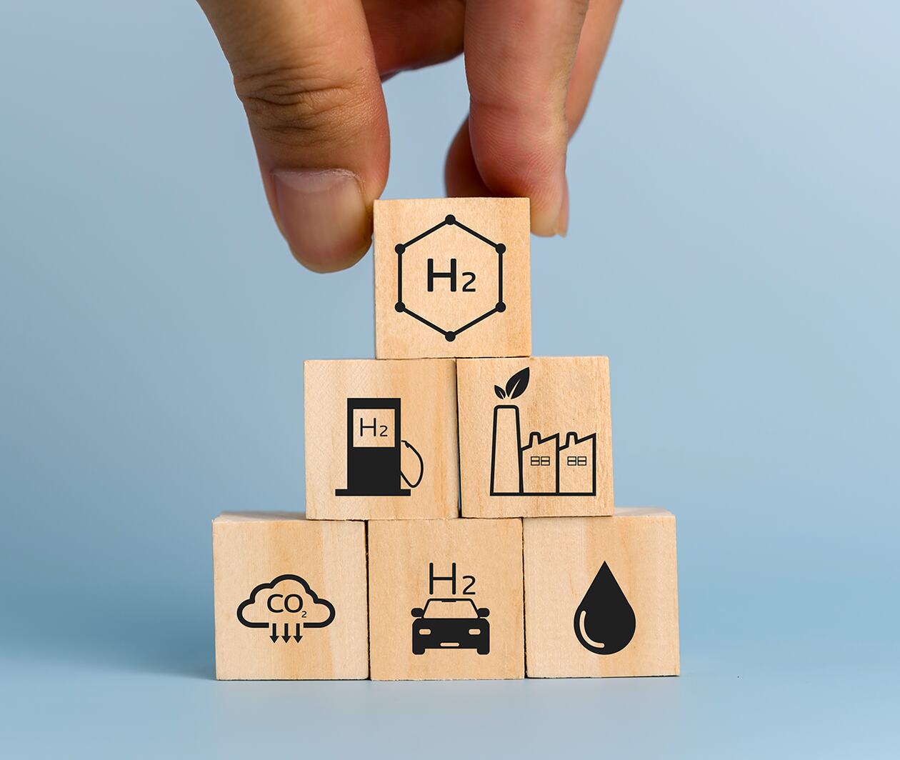 Fingers holding a wooden cube on top of a pile of five other cubes with various hydrogen related symbols.