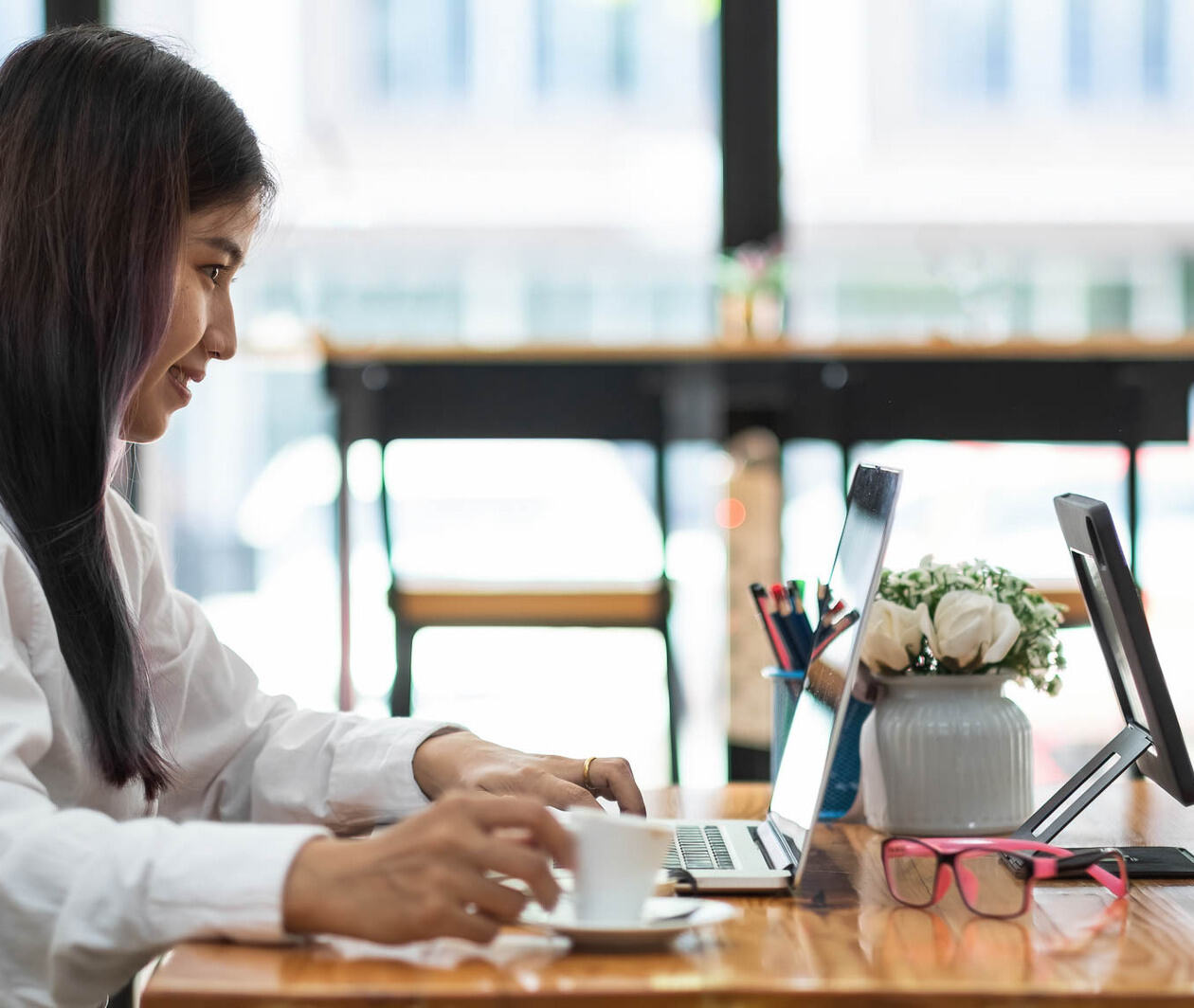 Confident young businesswoman using a laptop with a coffee. She is sat at a shared desk in front of large windows.