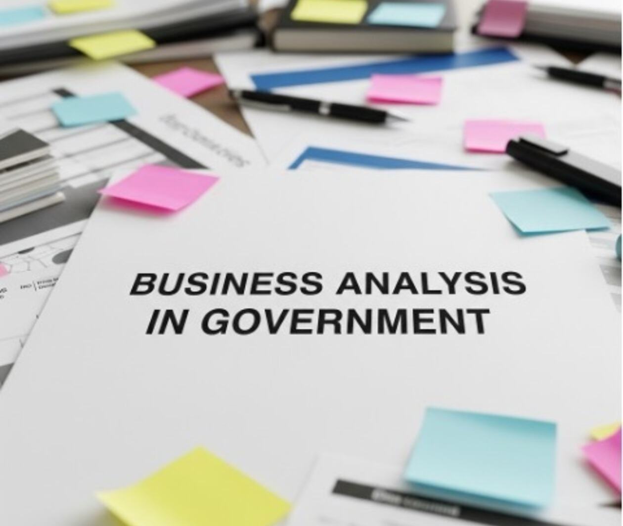 A close up of a desk covered in documents and post-it notes. A sheet of paper in the middle says "Business Analysis in Government"