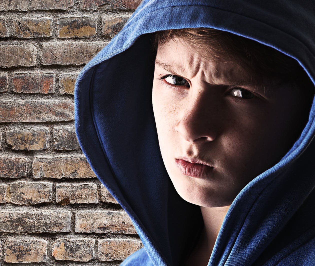 Boy in a hooded shirt scowling in front of a brick wall.