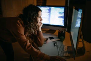 Young IT support woman using 2 monitors computer with coding document in a dark room, doing business in Web3. 