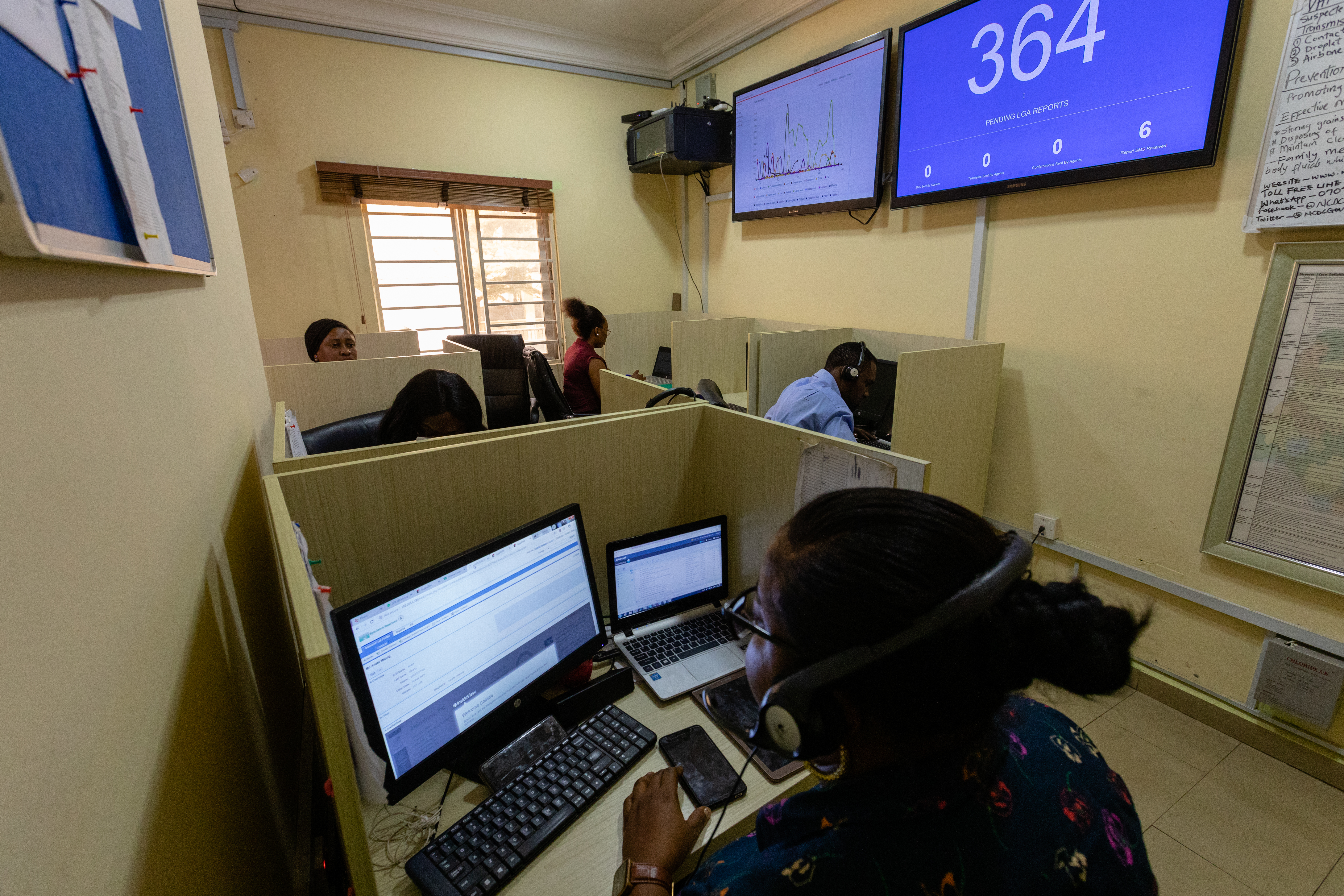 Five people sitting at their walled desks in a room, looking at computer screens. Two have headsets. There are three boards with written or printed notes. There are two full sized TV monitors, one displaying a coloured graph the other has the number 364.