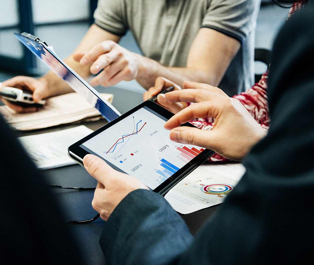 A person with a tablet with charts on screen. Someone else pointing at a clipboard.