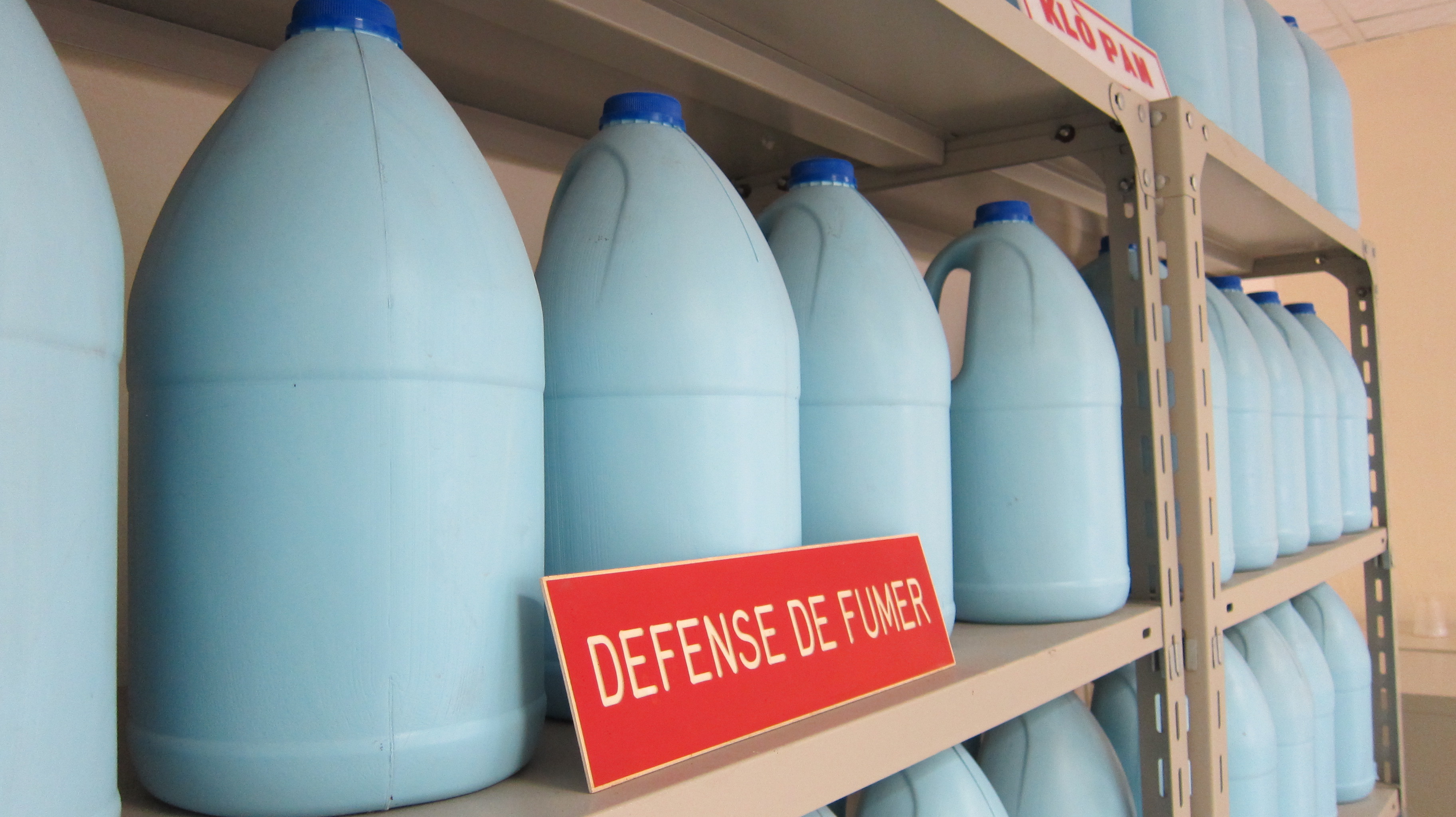 Four light blue bottles with dark blue caps of bleach on a metal shelf with a red sign that says, “Defense de fumer”. In the background there are more metal shelves with blue bottles.