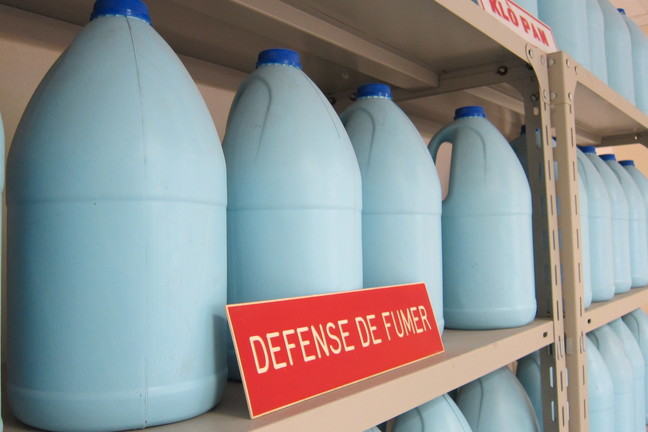 Four light blue bottles with dark blue caps of bleach on a metal shelf with a red sign that says, “Defense de fumer”. In the background there are more metal shelves with blue bottles.