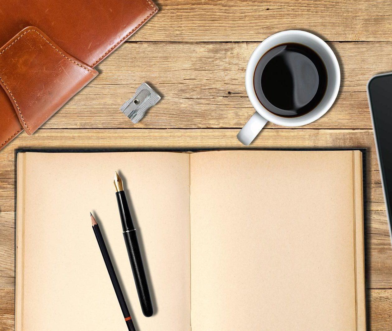 A pen and pencil sit on an open notebook with a coffee and an iPad close at hand, ready to start writing fiction