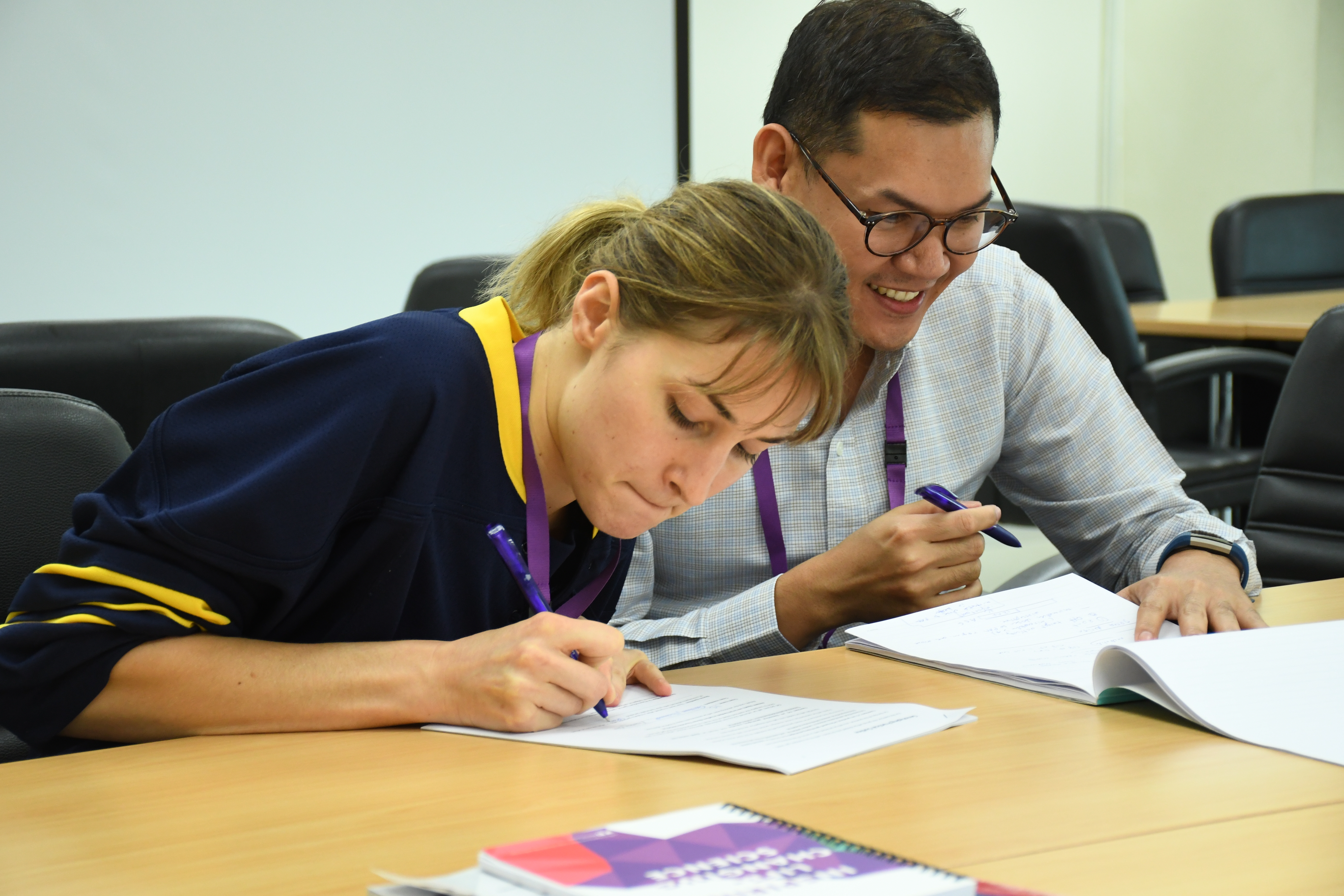 Learner in foreground concentrating on writing on a piece of paper and another learners in background smiling and holding a pen