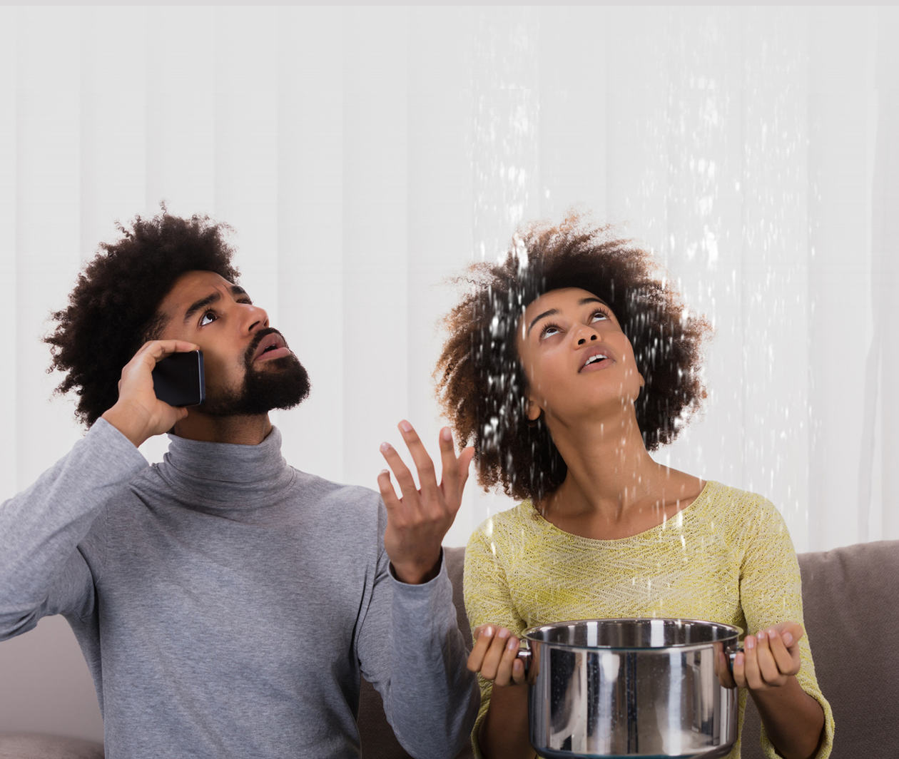 A couple are sitting on a sofa. The woman is using a large pan to catch water that is coming from the ceiling. The man is on the telephone calling their personal lines insurer to make a claim on their household insurance policy.