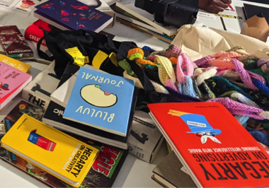 A selection of creative books stacked on a table, there is also a multicoloured crochet chain, black tote bag, and white tote bag on the table.