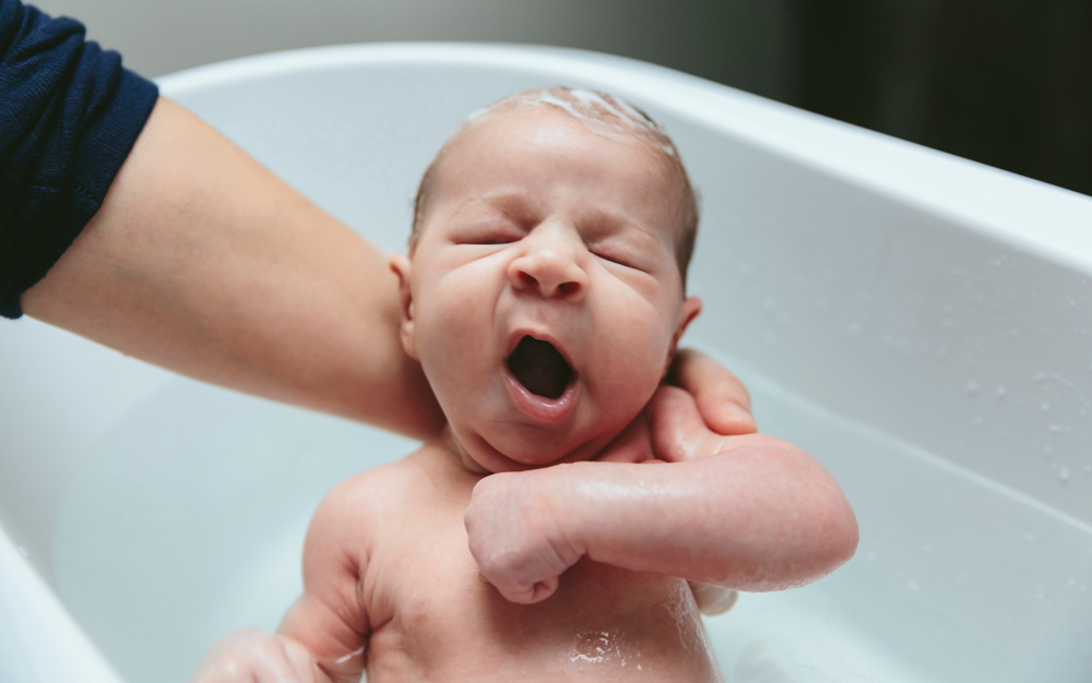 A newborn baby yawns as he is being bathed in the tub.