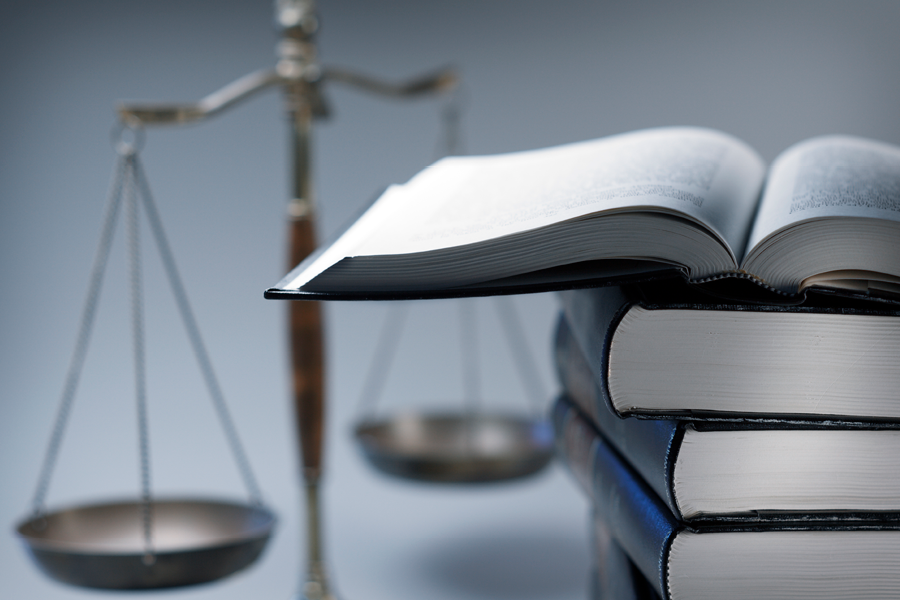 A stack of law books stands in front of a justice scale that is slightly out of focus. On top of the stack is an open law book.