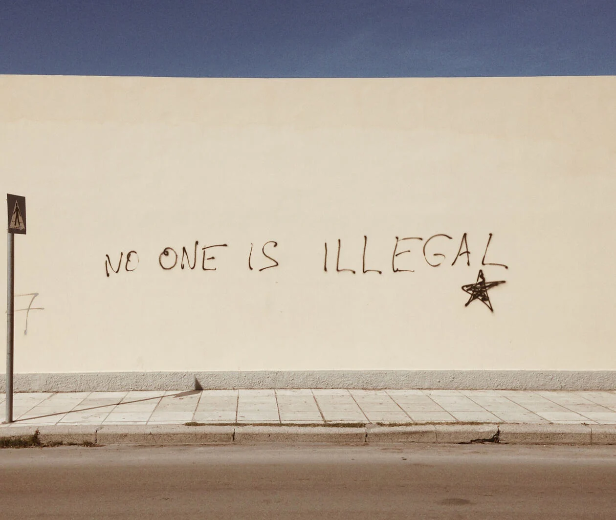 A cream wall beside a street with the graffiti stating "No one is illegal"