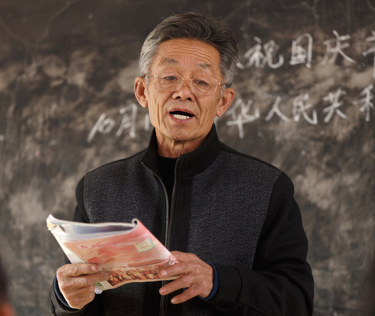 Elderly male teacher holding book with Chinese text on black board behind him.