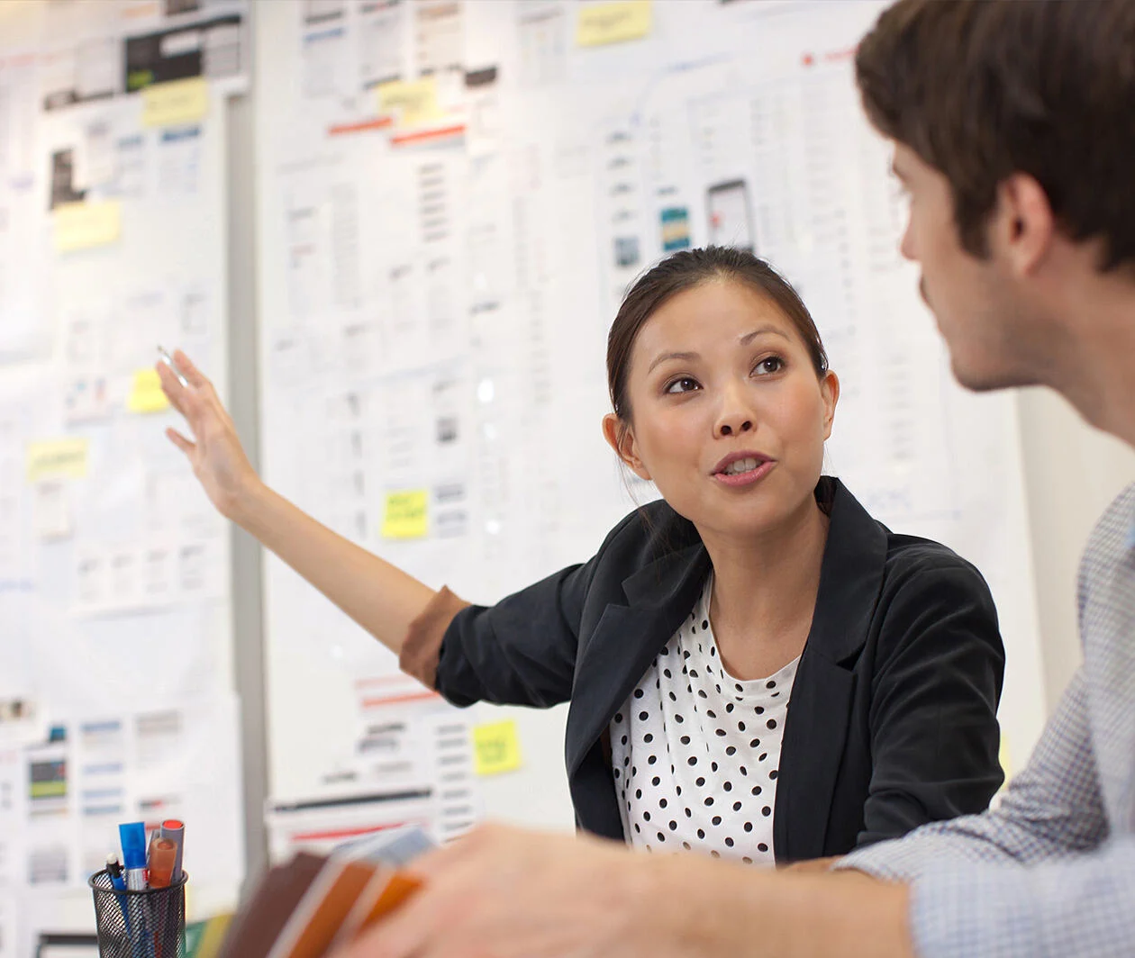 Woman talking to a man pointing at a wall of posters and sticky notes behind her.