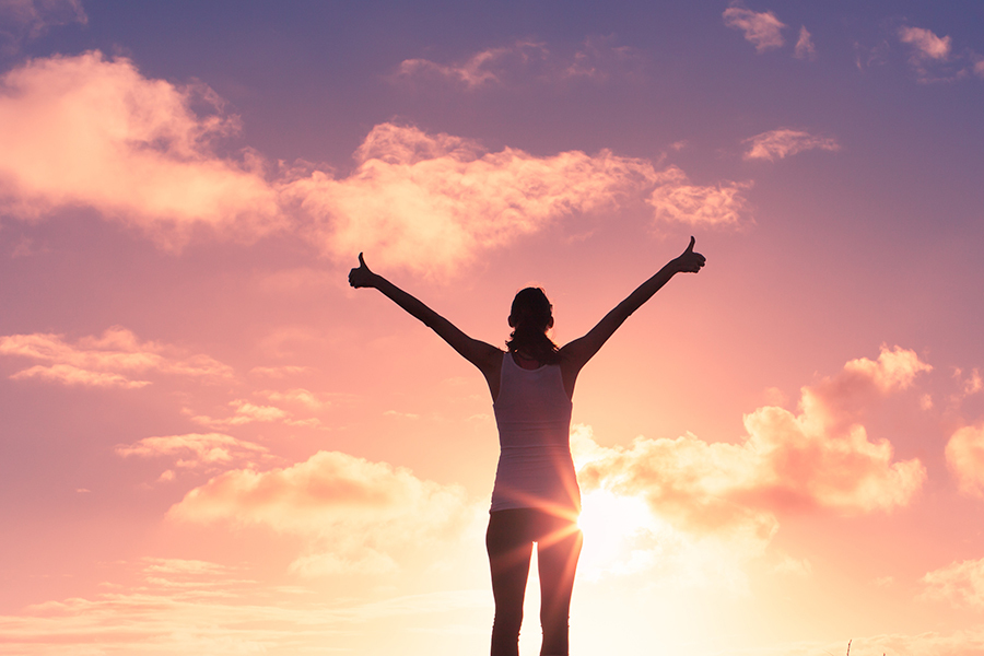 A woman standing in front of a beautiful sunset giving two thumbs up.