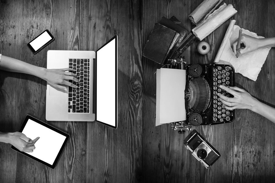 Two users at wooden table, one with laptop, tablet and smartphone, the other with typewriter, notebook and camera