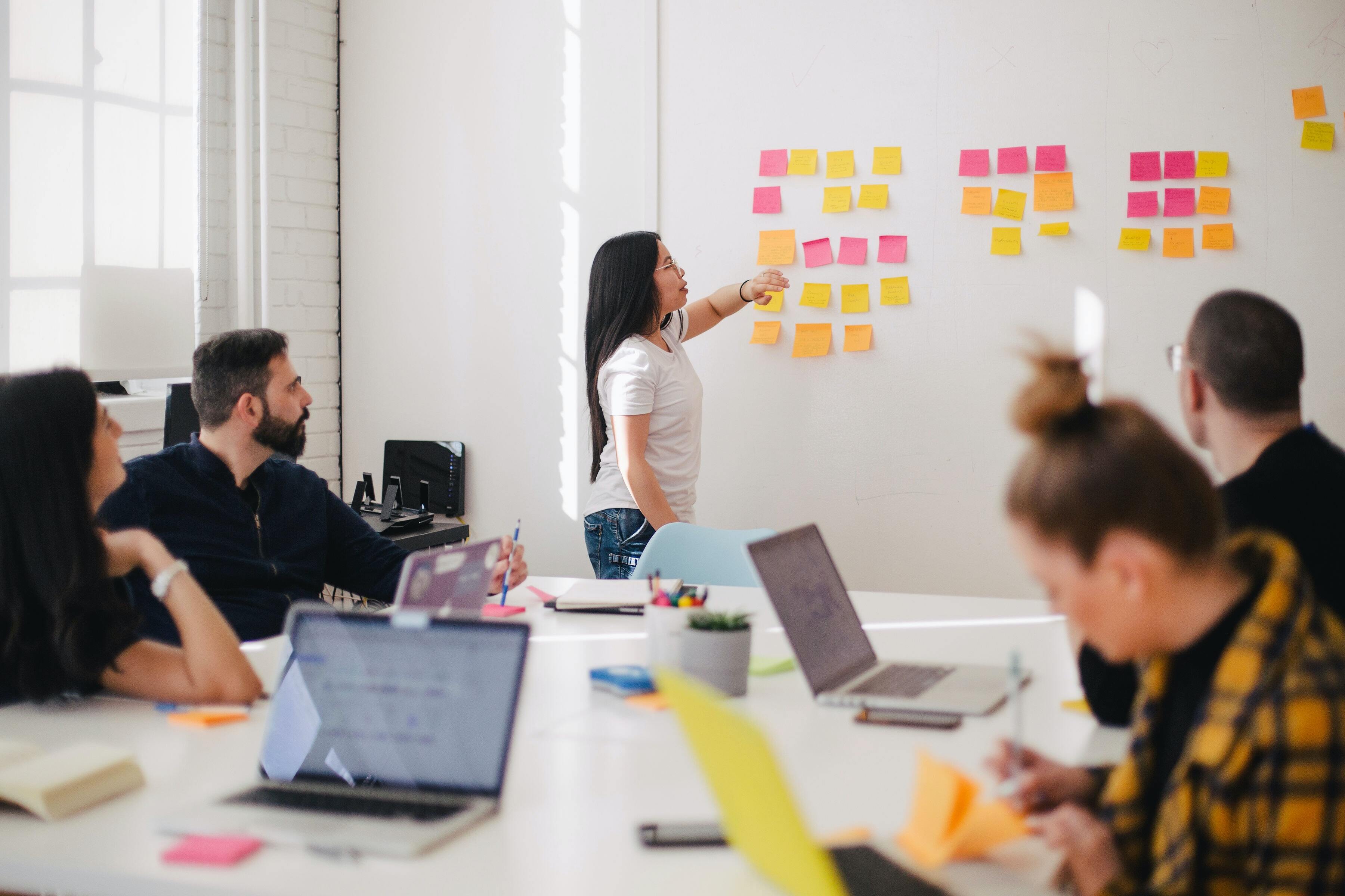 A boardroom with people sitting around a table and a woman sticking postit notes on the wall