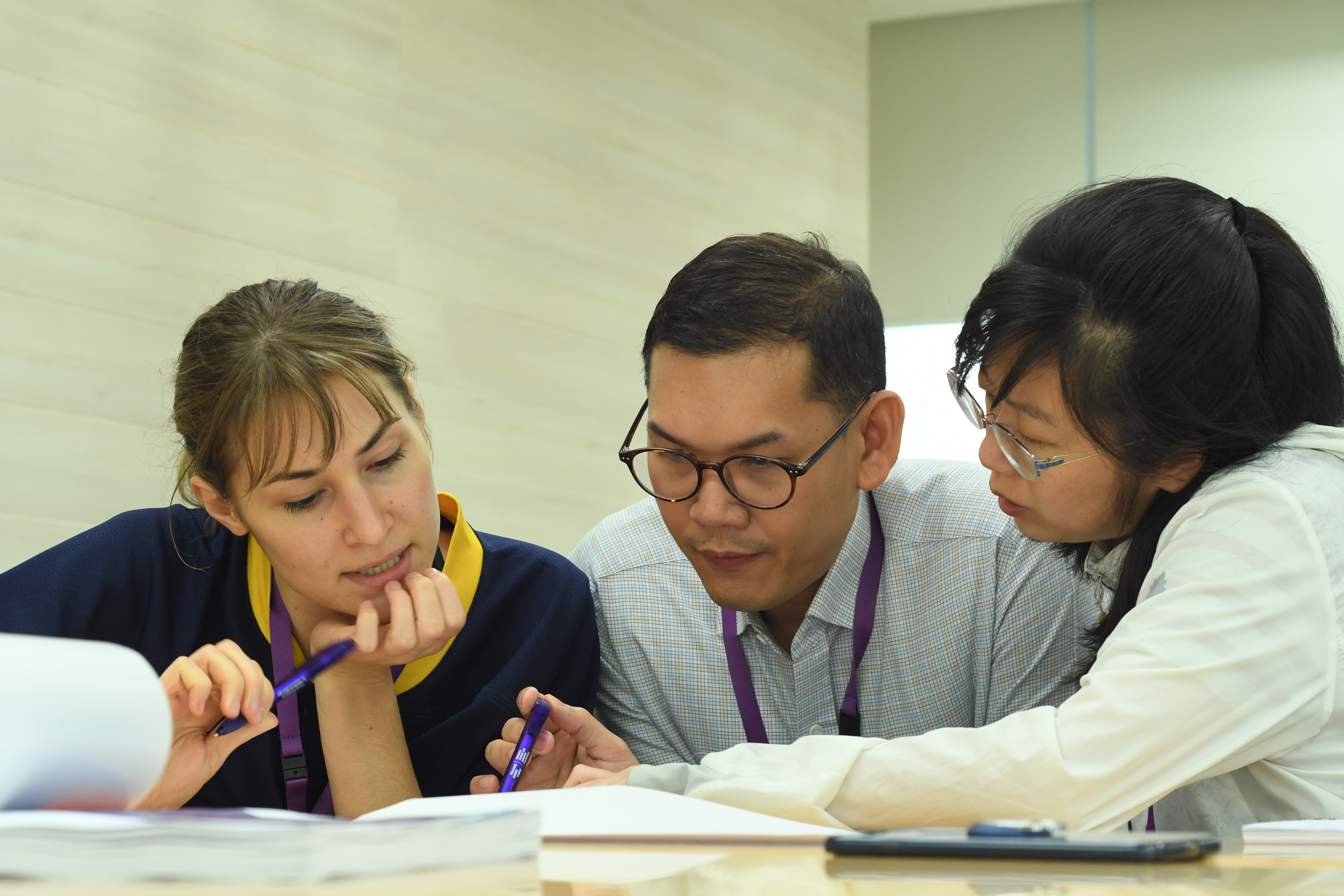 three learners sitting at a desk and concentrating on some work involving writing, one of them pointing something to others