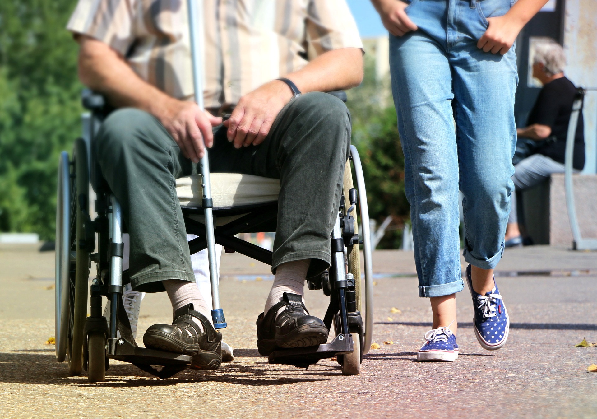 Photo of two people's legs as they move along a pavement. An older person in aa wheelchair and a teenager walking at their side.