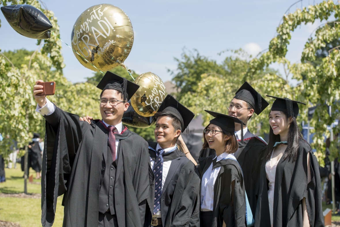 The image shows a group of graduates at their graduation ceremony stood outside in the sunshine smiling and taking a group selfie together.