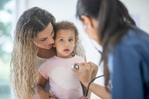 A mother holds her daughter while a doctor checks the child with a stethoscope