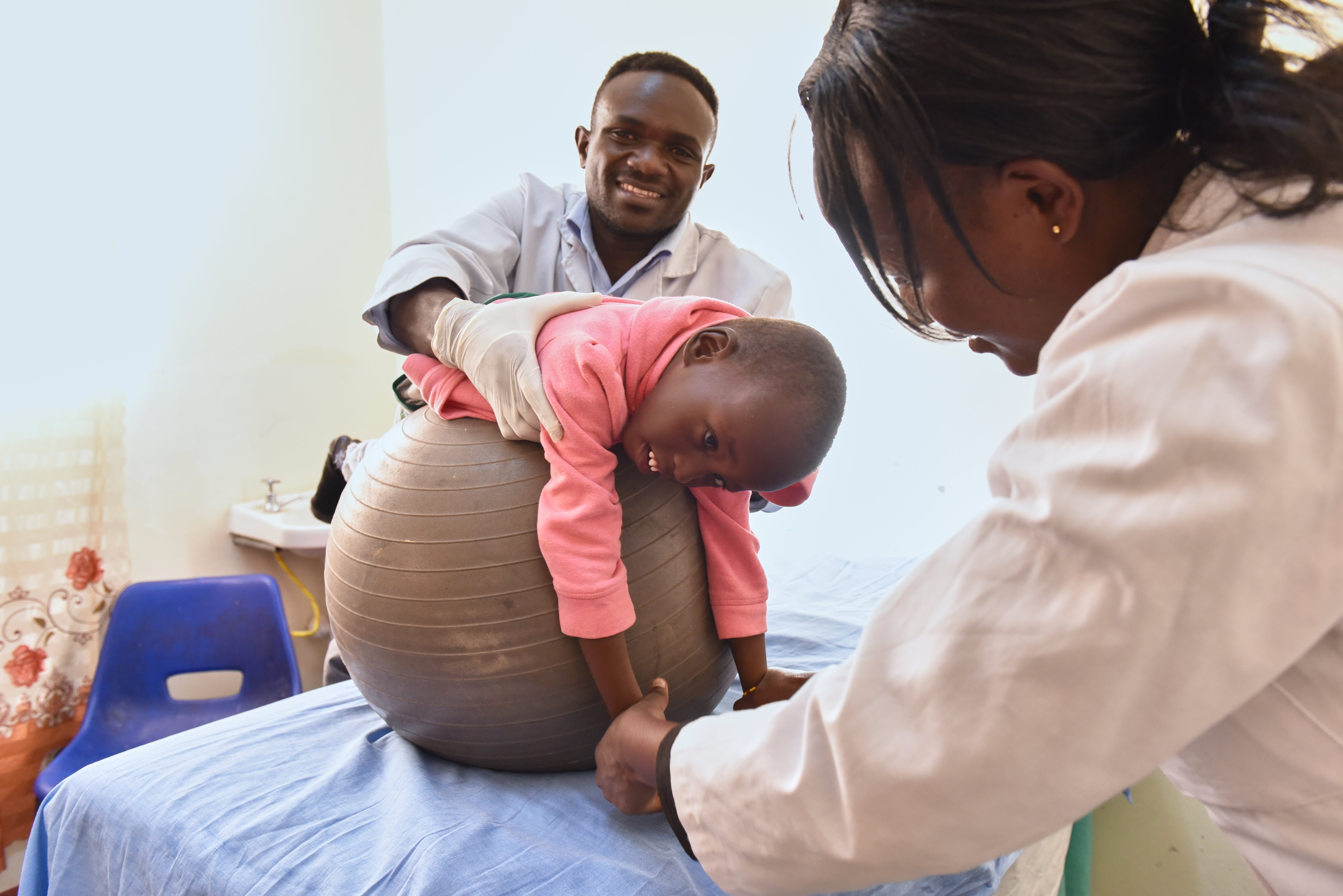 Two healthcare professionals support a boy through physiotherapy. The boy is being supported as he lies on a large exercise ball.