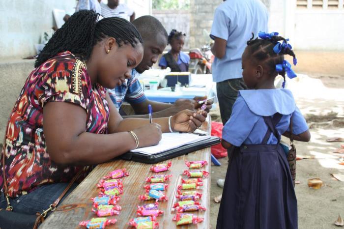 This image depicts a Haitian man and woman sat at a table. The woman is filling out a paper form with a pen. In front of the table, standing, is a small girl wearing a blue school uniform. There are many individually wrapped sweets on the table.