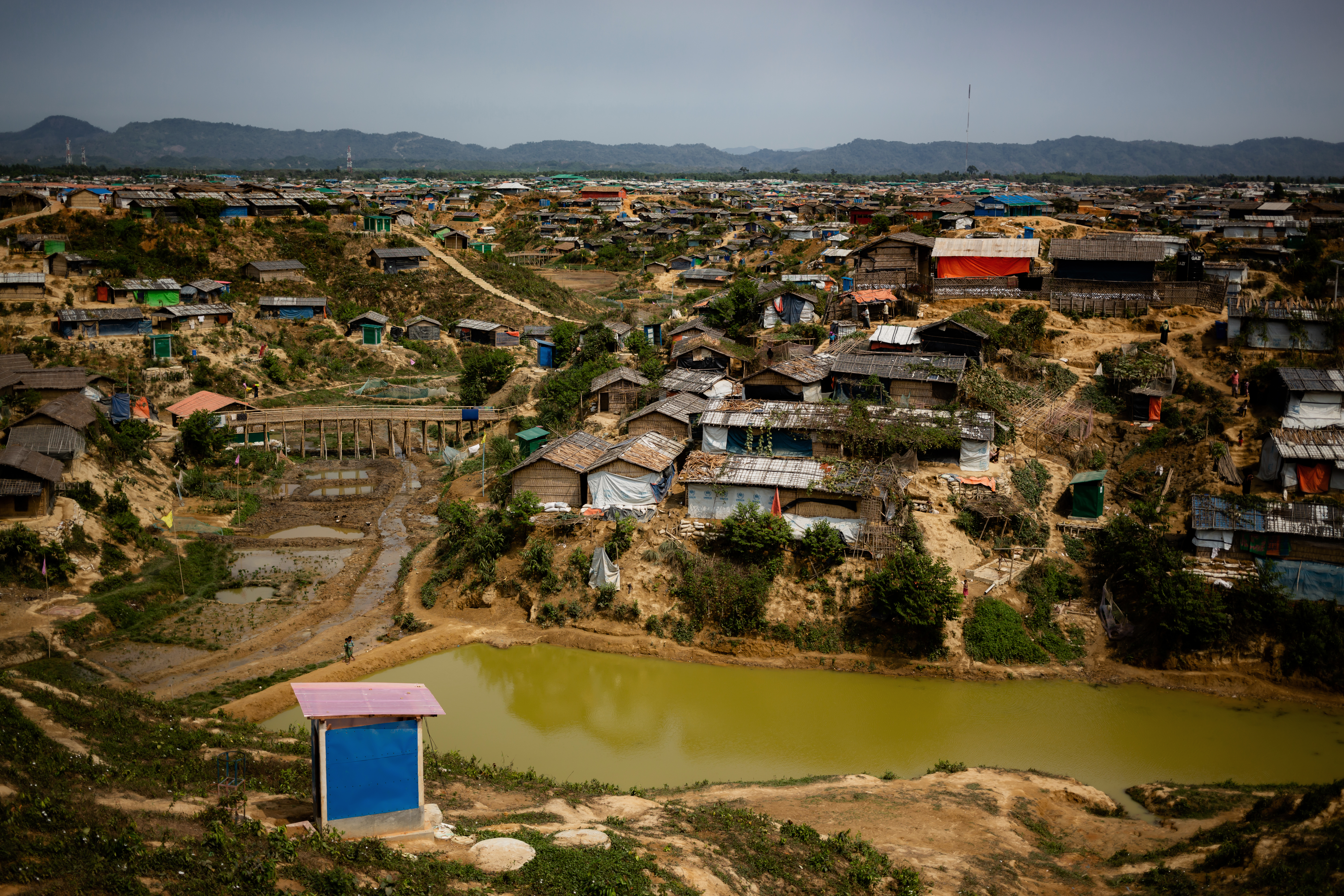 The Kutapalong Rohingya refugee camp is composed of hundreds of houses built on a muddy, piece of land with a river flowing in front of it. A shed stands on the opposite side of the river. This is home to more than one million displaced people.