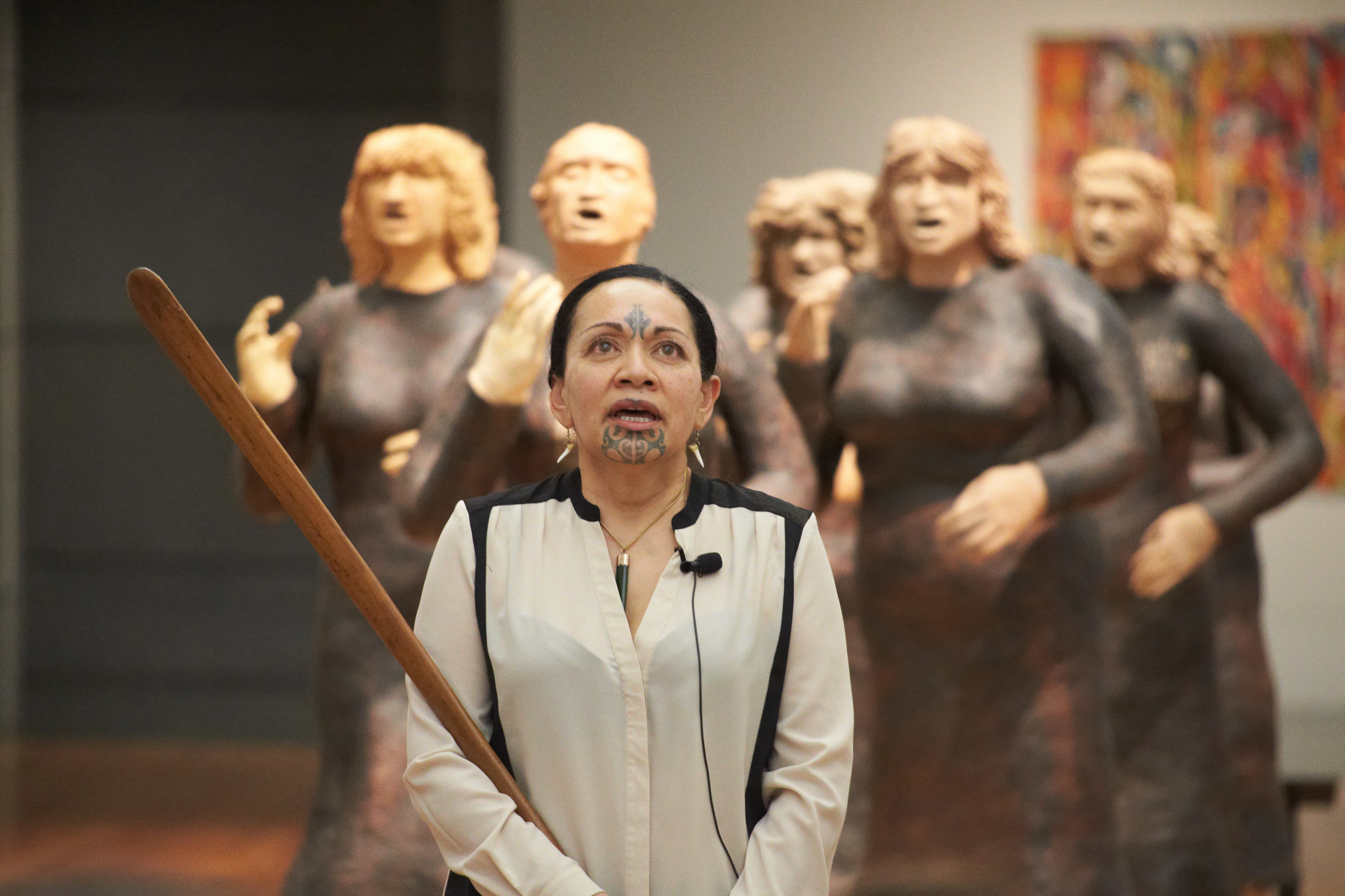 A Māori woman with traditional facial tattoos and holding a ceremonial weapon sings in front of an exhibition featuring the figures of Māori woman.