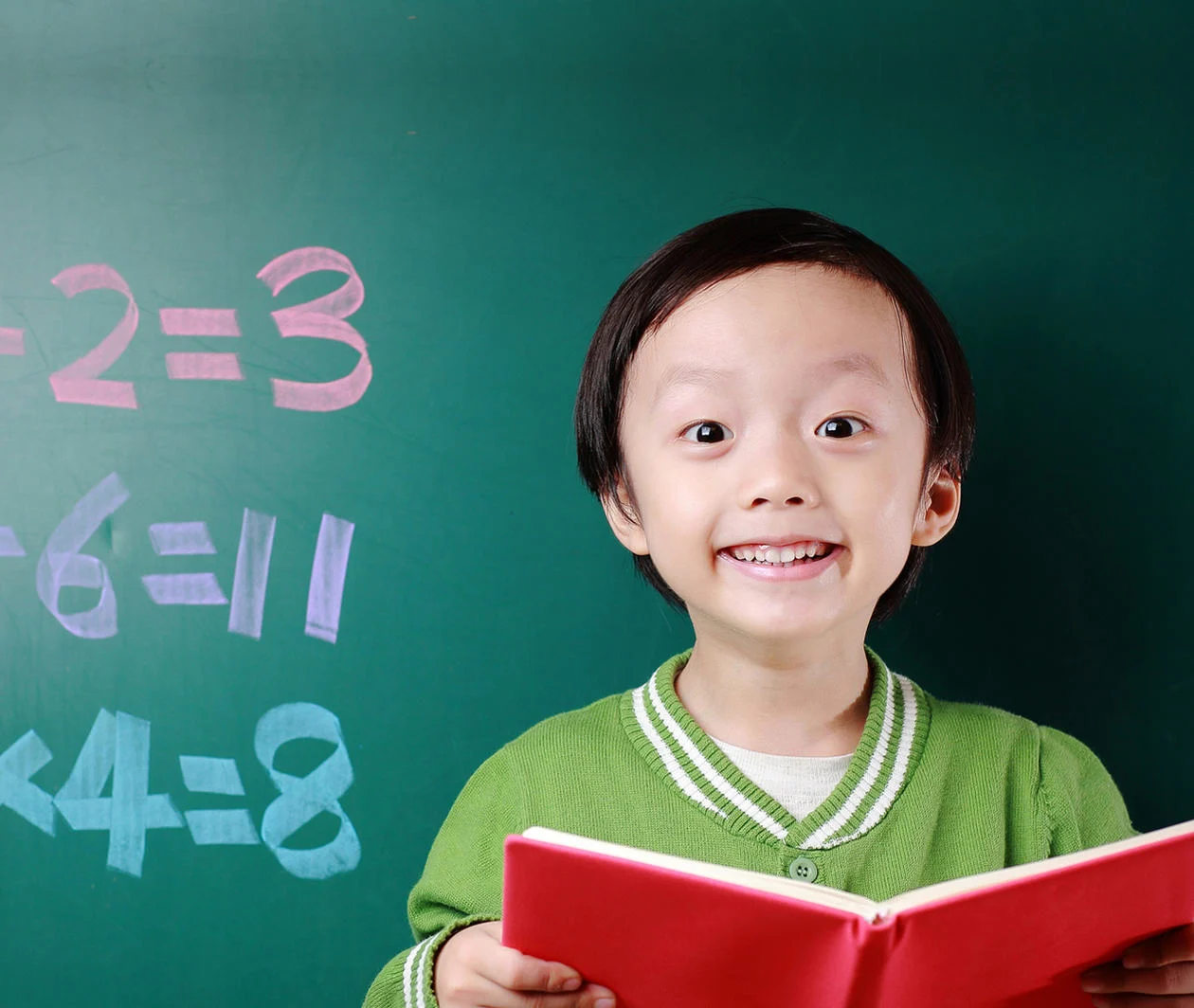 A child holds an open book in front of a blackboard