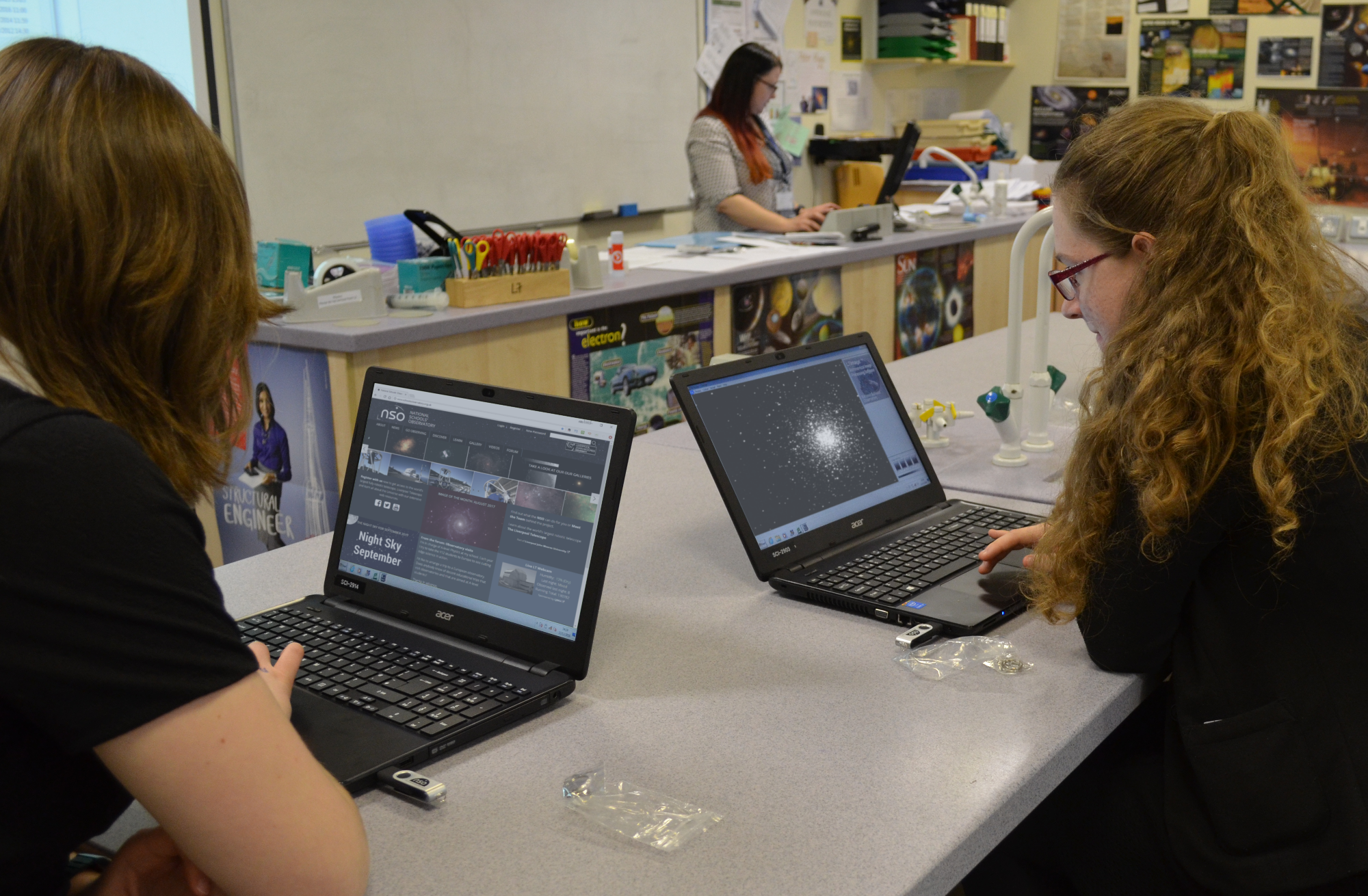 Two female students working on their laptops in a classroom, looking at the National School's Observatory website