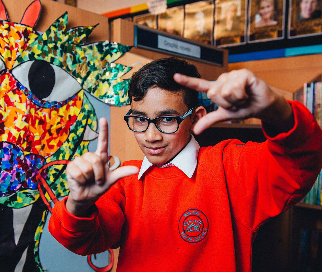 A young boy in school uniform holds his hands up in the shape of a camera viewfinder as if framing a film shot. 