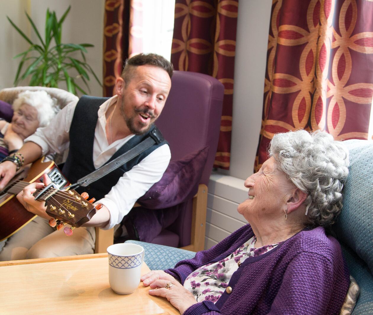 A musician singing with a guitar in a care home and an elderly woman smiling sitting next to him.