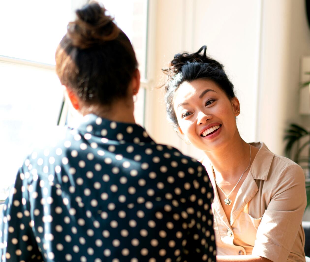 Two women seated at table talking. One has back to camera with hair in bun. Other is facing her, listening and smiling. 