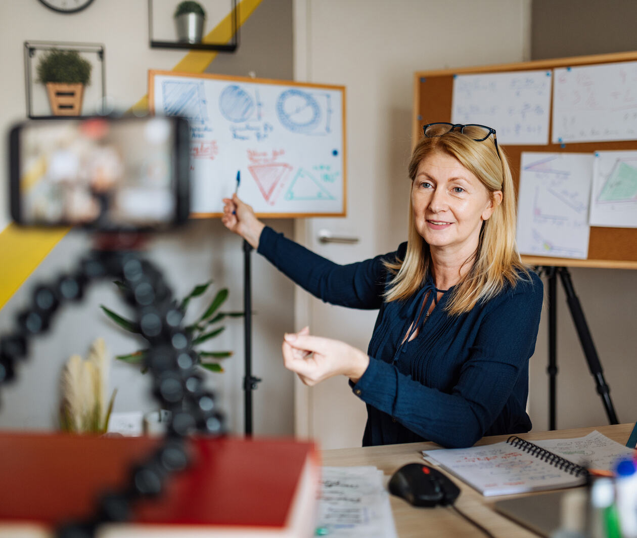 A teacher conducting a class from her home, recording it through her smartphone. There are whiteboards and cork boards behind her which she is pointing to