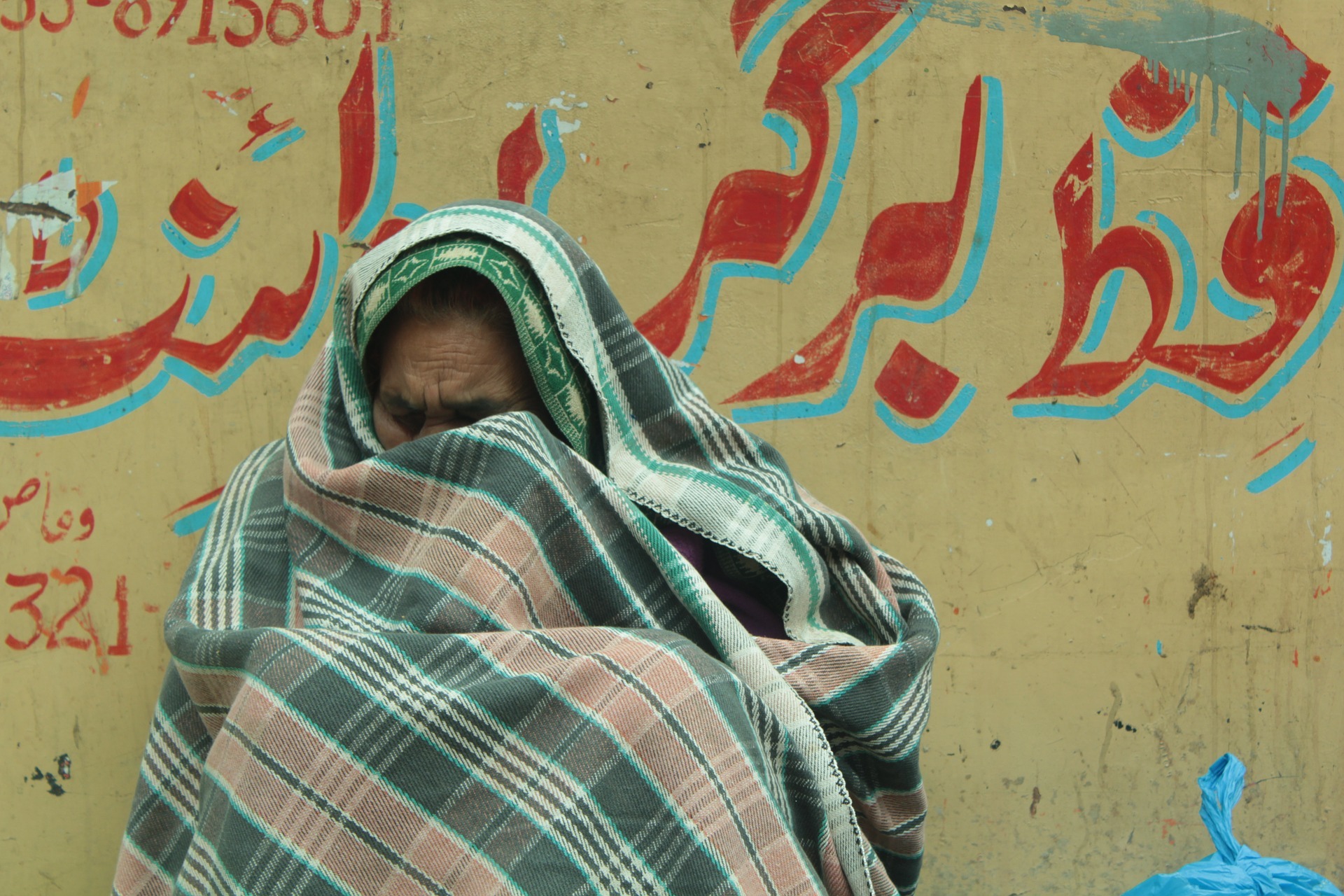 Older refugee sitting against a wall wrapped in a blanket.