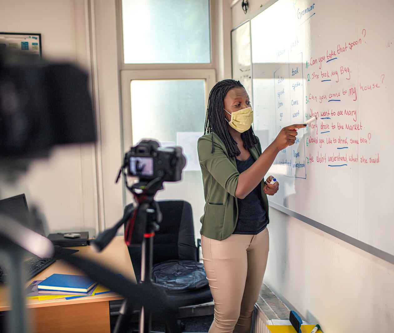 A teacher with a face mask on, standing at a full size whiteboard, underlining sentences with a dry marker. In the foreground are two cameras which she is presenting to