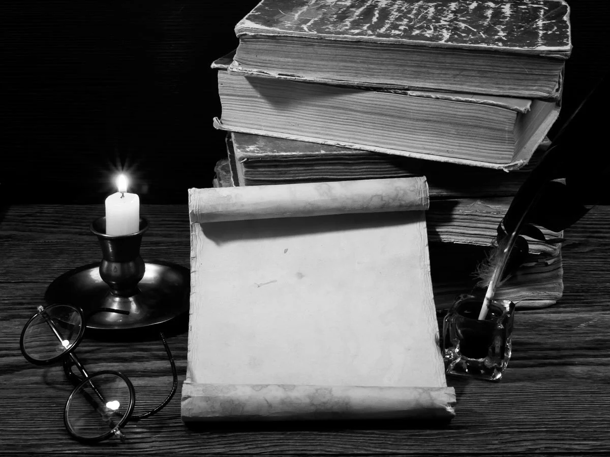 Pile of books on a table with quill and ink bottle, parchment, candle and reading spectacles