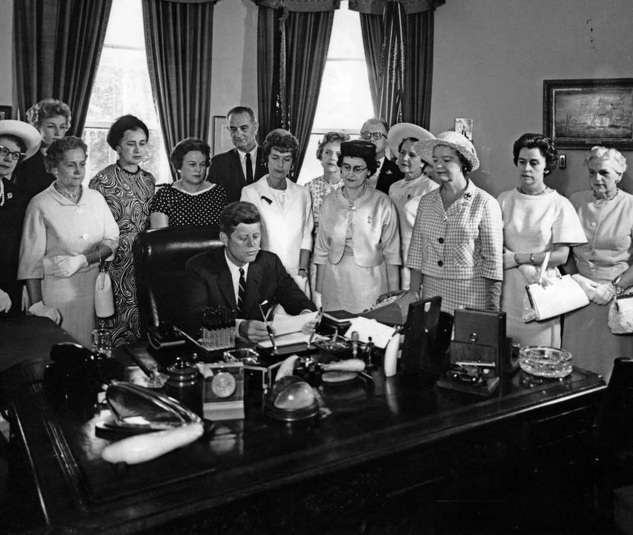 Women surrounding President JFK in the Oval Office.