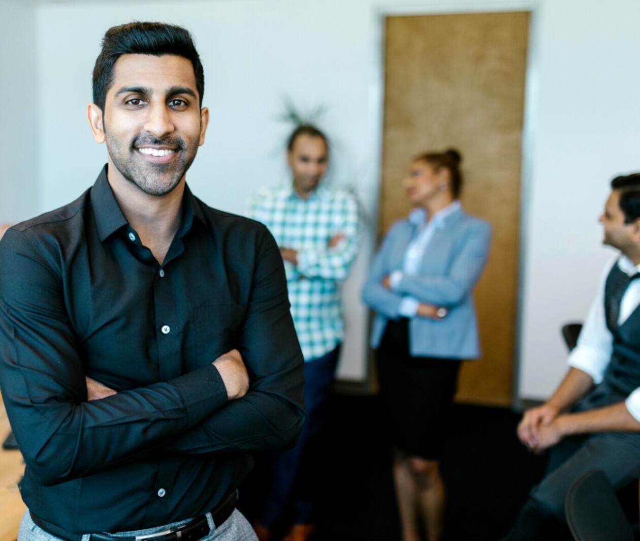 A man cross his arms smiling and standing in front of his colleagues chatting at the background, slightly blurred