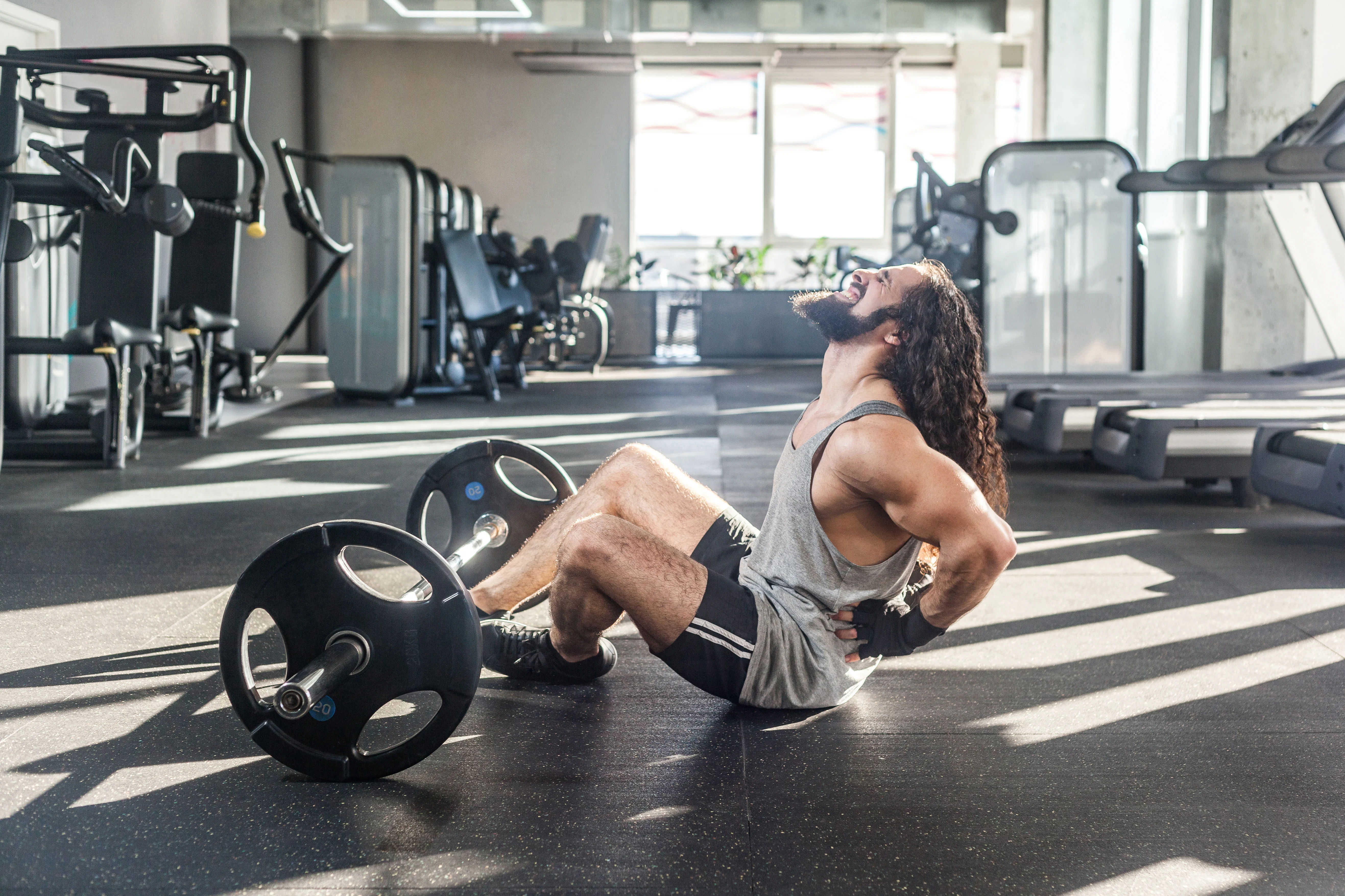 Portrait of injury young adult man athlete with long curly hair working out in gym, sitting on floor and have strong hurt problem with back, spasm painful