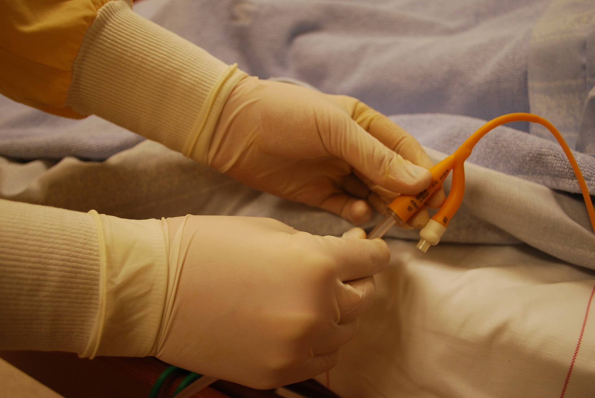 Image depicting gloved hands holding a catheter by a hospital bed