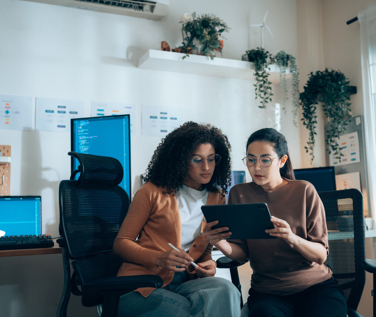Two women looking at a tablet with computer screens behind them